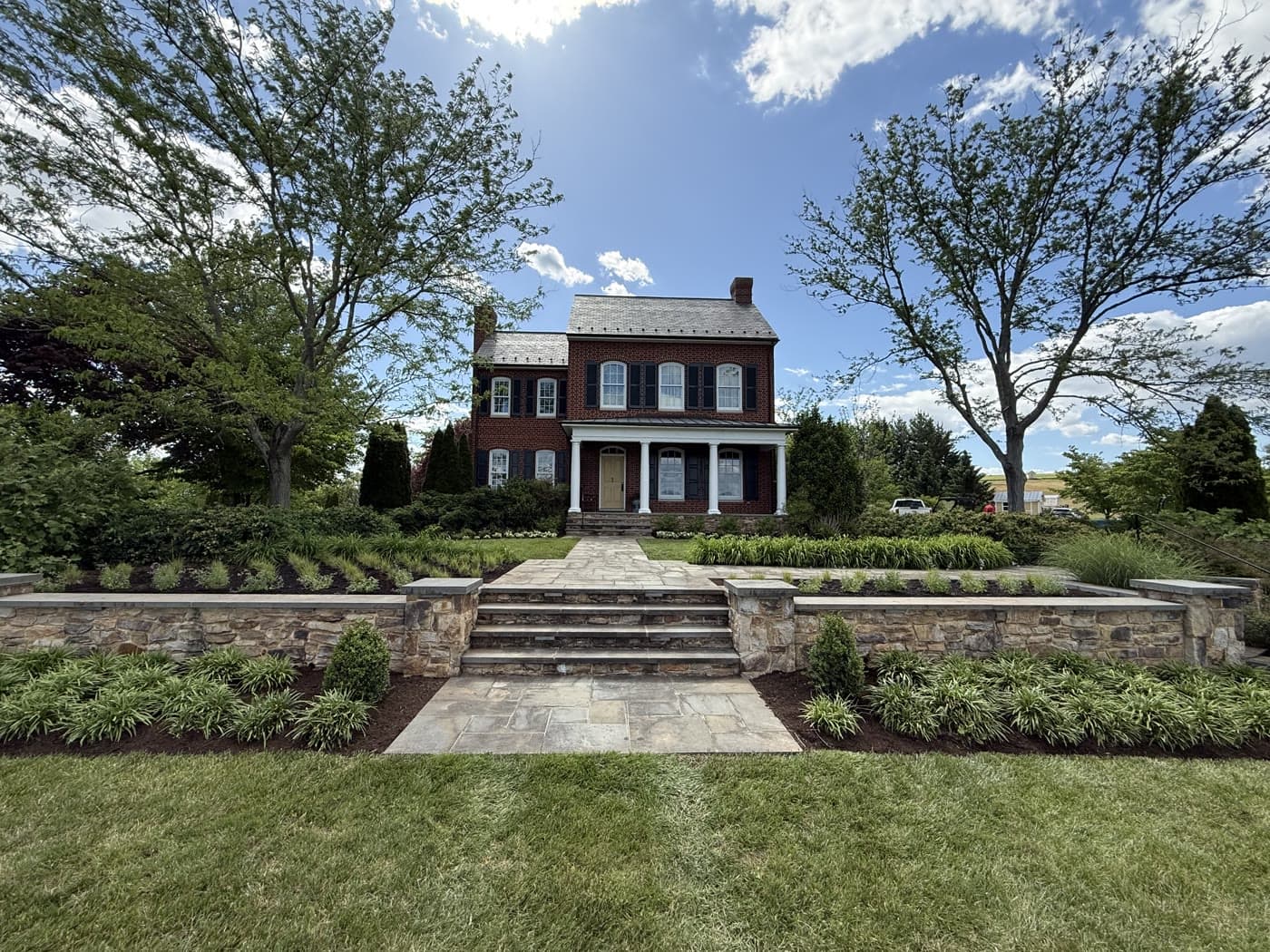 Wide view of the finished front landscape: stone terraces, planted beds, and the brick house